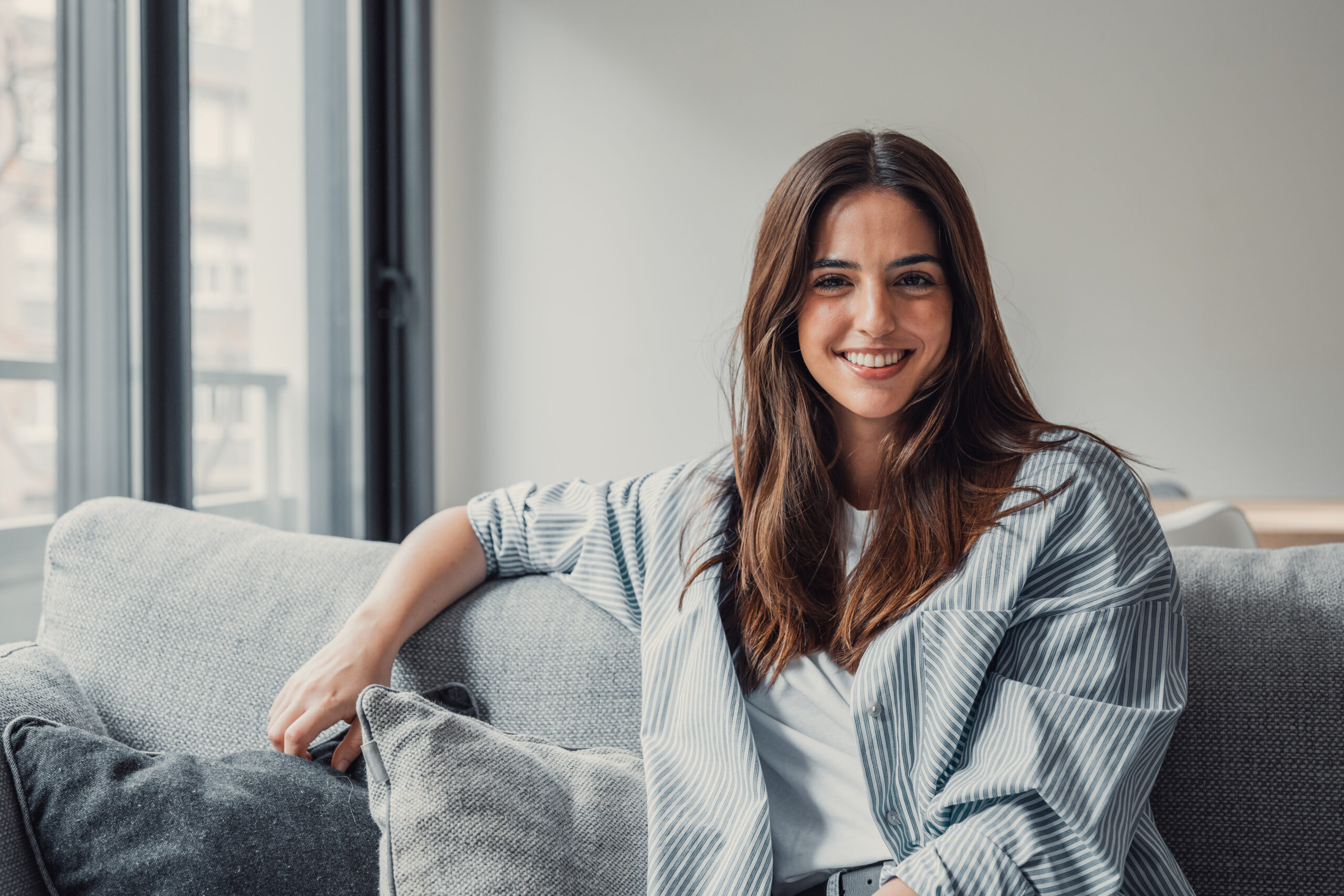 Happy attractive young adult woman girl sitting on sofa looking at camera in modern cozy home, smiling single 20s lady beautiful face relaxing on couch alone enjoy wellbeing posing for portrait. How Cellular Rejuvenation Supports Healthier, More Youthful-Looking Skin